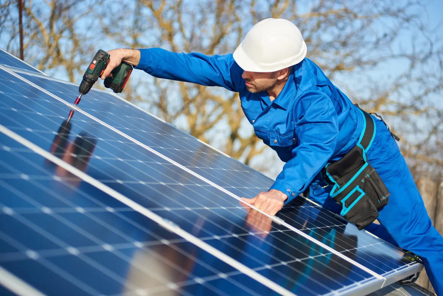 Arbeiter in blauem Anzug und Schutzhelm bei der Installation einer Photovoltaikanlage mit Schraubenzieher. Elektriker montiert Modul auf dem Dach eines modernen Hauses. Alternative Energie ökologisches Konzept.